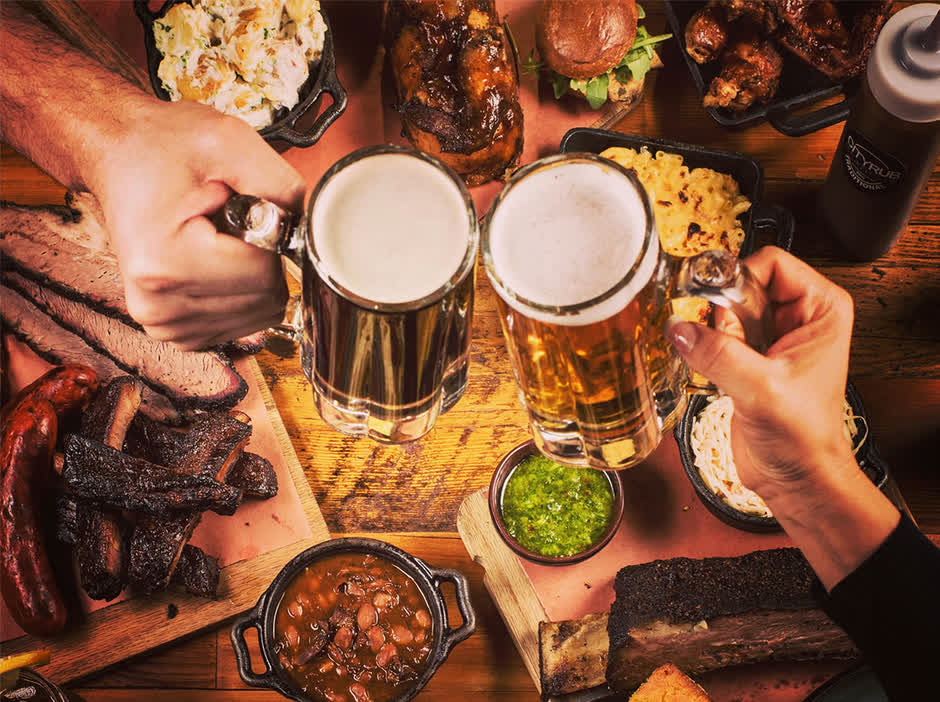 A table of City Rub food from above — brisket, ribs, sausage, mac and cheese, beans and cornbread — with two beers being clinked over it.